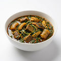 A white ceramic bowl filled with a rich, dark stew containing chunks of meat, yellowish-brown roots, and abundant green leafy vegetables, set against a clean white background.