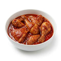 A white ceramic bowl filled with cooked chicken drumsticks and thighs in a rich, glossy red tomato-based sauce, set against a clean white background.