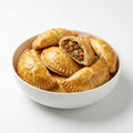 A white bowl filled with golden-brown baked hand pies, with one pie cut open to show a savory meat and vegetable filling, photographed on a clean white background.